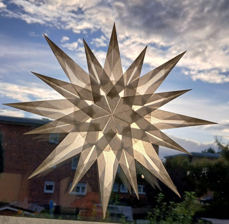 May include: A decorative star, made from translucent paper, attached to a window. The star has multiple points and a geometric design. The background shows a cloudy sky and a building. The star's design allows light to pass through.
