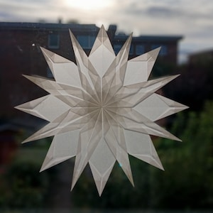 May include: A large, white paper star decoration with multiple points, displayed on a window. The star is made of folded paper, creating a layered, geometric design. The background shows a blurred view of a building and trees.