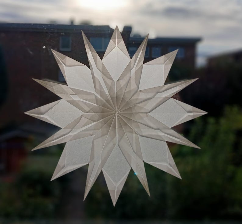May include: A large, white paper star decoration with multiple points, displayed on a window. The star is made of folded paper, creating a layered, geometric design. The background shows a blurred view of a building and trees.