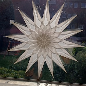 May include: A decorative star, made from a translucent, off-white material, with a geometric design and multiple points. The star is attached to a window, with a view of a garden and a building visible through the glass. The star's design creates a layered effect.