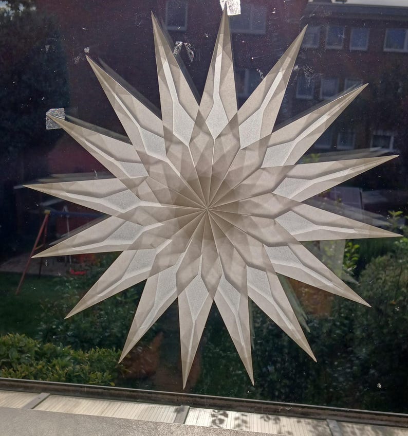 May include: A decorative star, made from a translucent, off-white material, with a geometric design and multiple points. The star is attached to a window, with a view of a garden and a building visible through the glass. The star's design creates a layered effect.