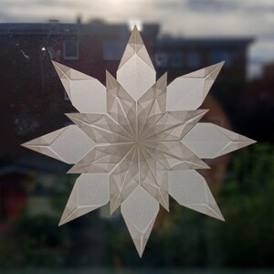 May include: A large, geometric paper star decoration with a layered design. The star is made of translucent, off-white paper, with sharp, angular points and a central, circular pattern. The star is displayed against a blurred background of a building and sky.