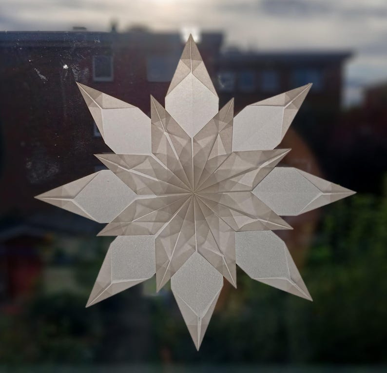 May include: A large, geometric paper star decoration with a layered design. The star is made of translucent, off-white paper, with sharp, angular points and a central, circular pattern. The star is displayed against a blurred background of a building and sky.