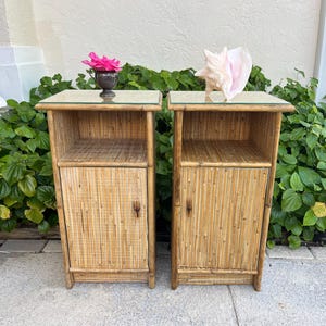 May include: A pair of matching rectangular bedside tables constructed from light brown bamboo and rattan. Each table features an open top shelf, a single cabinet door with a small wooden handle, and a flat glass top surface for a clean finish.