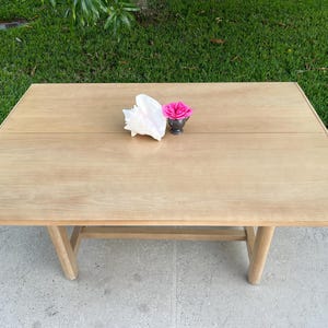 May include: A light-colored wooden table with a seashell and a small silver container holding a pink rose. The table has a rectangular top and angled legs. The table is set outside on a concrete surface.