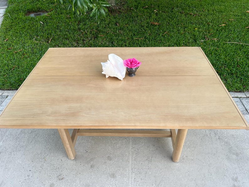 May include: A light-colored wooden table with a seashell and a small silver container holding a pink rose. The table has a rectangular top and angled legs. The table is set outside on a concrete surface.