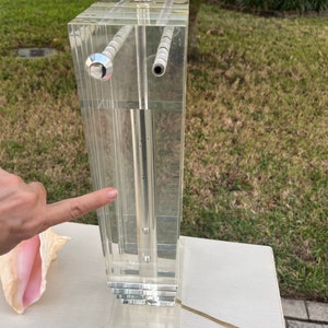 May include: Clear acrylic table lamp with a rectangular design. The lamp features a silver-colored metal base and a clear cord. The lamp is composed of multiple clear acrylic panels. The lamp is on a white surface.