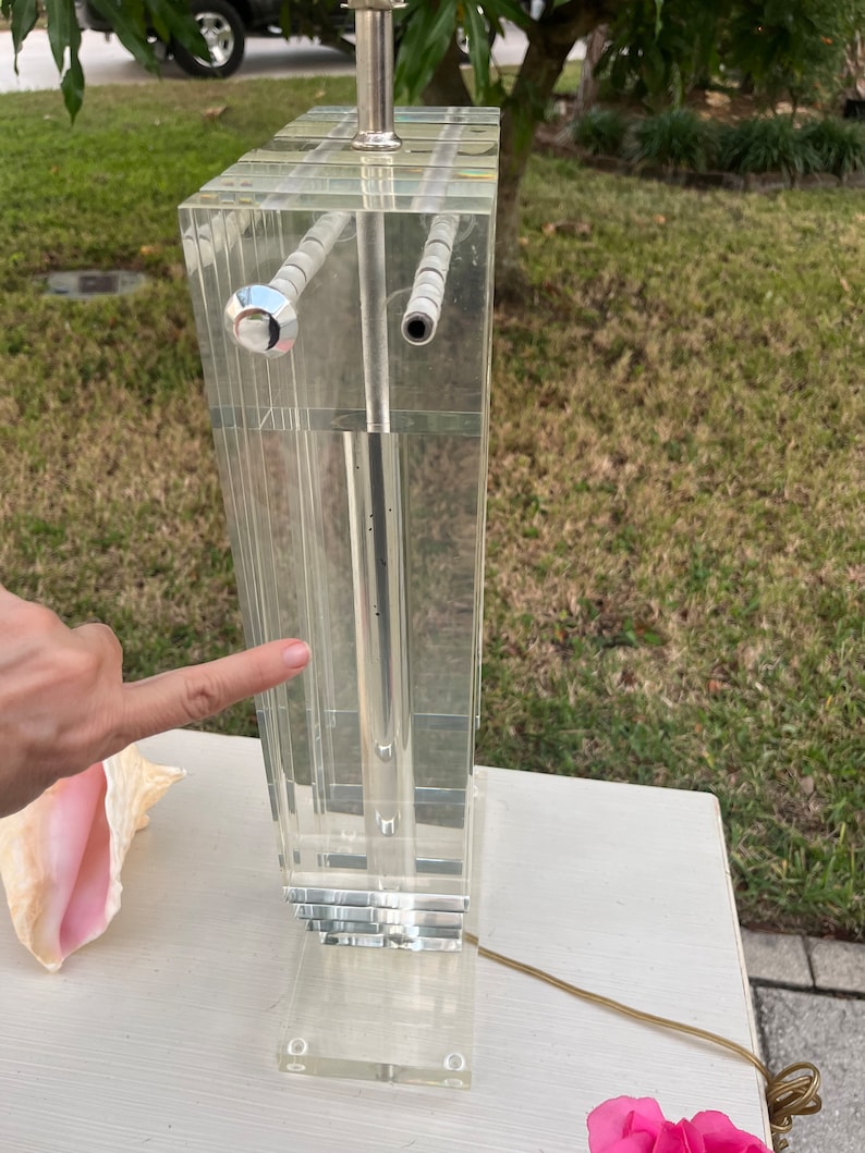 May include: Clear acrylic table lamp with a rectangular design. The lamp features a silver-colored metal base and a clear cord. The lamp is composed of multiple clear acrylic panels. The lamp is on a white surface.