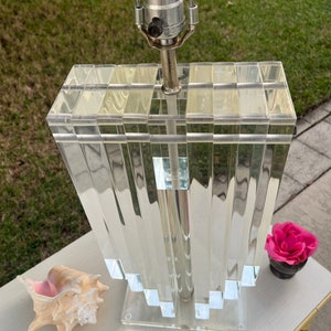 May include: A clear acrylic table lamp with a geometric design. The lamp has a silver metal neck and a clear base. A seashell and a small pot with a pink flower are next to the lamp.
