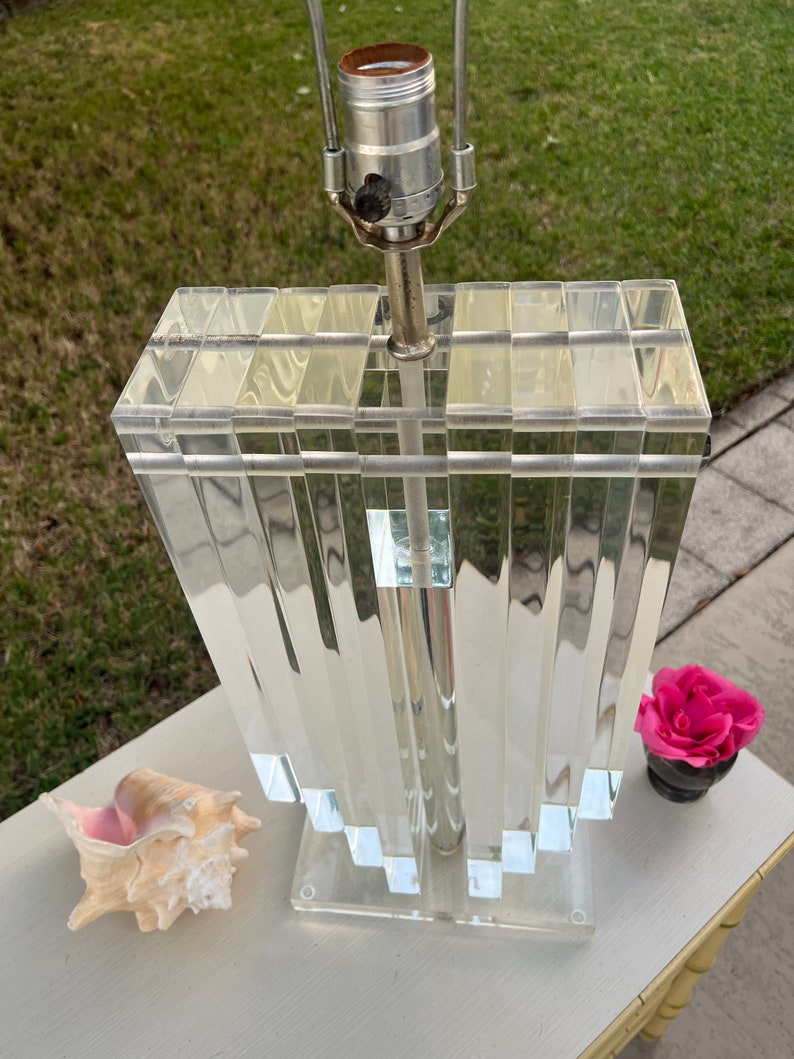 May include: A clear acrylic table lamp with a geometric design. The lamp has a silver metal neck and a clear base. A seashell and a small pot with a pink flower are next to the lamp.