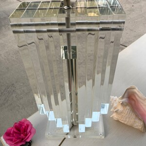 May include: Clear acrylic table lamp with a silver metal base. The lamp features a rectangular shade with vertical, ridged panels. A pink rose and a seashell are next to the lamp on a white surface.