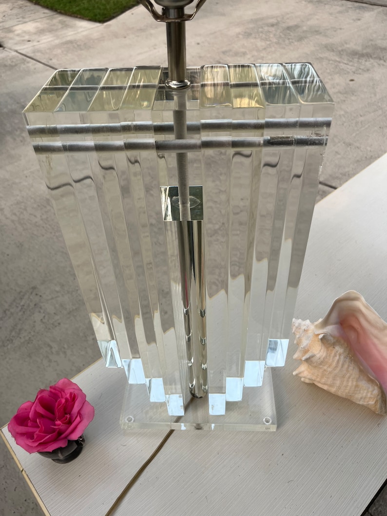 May include: Clear acrylic table lamp with a silver metal base. The lamp features a rectangular shade with vertical, ridged panels. A pink rose and a seashell are next to the lamp on a white surface.
