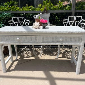 May include: A white wicker desk with two drawers and a woven top. The desk has a decorative edge and is supported by angled legs. A pink rose, seashell, and decorative items sit on top. Two white chairs are in the background.