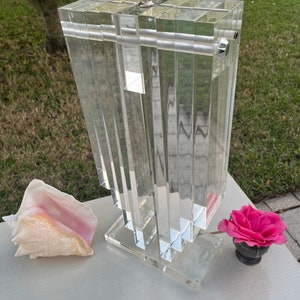 May include: A clear acrylic lamp with a rectangular, layered design. The lamp has a silver-colored metal base and a white lampshade. A conch shell and a pink rose in a small black vase are next to the lamp.