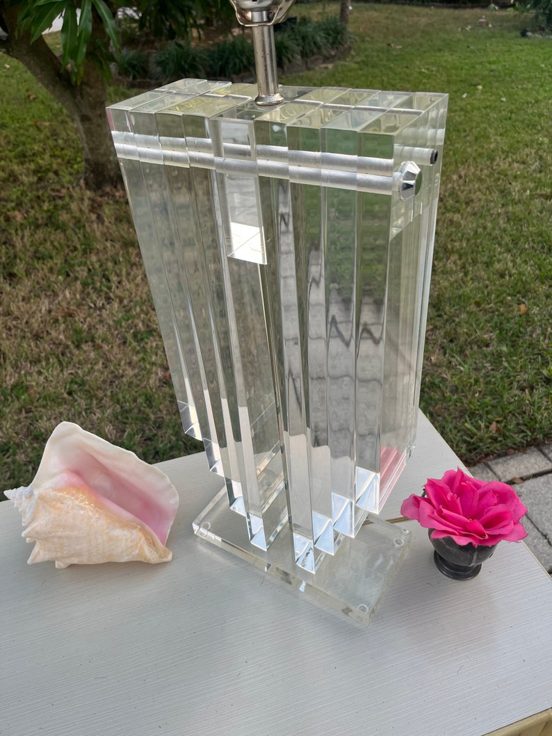 May include: A clear acrylic lamp with a rectangular, layered design. The lamp has a silver-colored metal base and a white lampshade. A conch shell and a pink rose in a small black vase are next to the lamp.