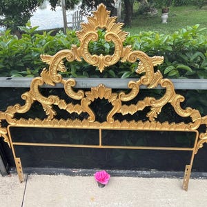 May include: Ornate gold-colored headboard with intricate scrollwork and a black upholstered center. The headboard has a decorative top and is supported by metal legs. A pink flower rests in front of the headboard.