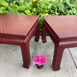 May include: Two square, red side tables with a woven texture on the tabletop and legs. A pink flower in a small black vase sits between the tables. The tables are made of wood and have a classic design.