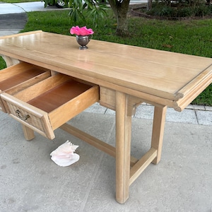 May include: A light-colored wooden console table with two drawers open. The drawers have woven rattan accents and ornate handles. A small silver vase with a pink flower sits on top. The table is approximately 60 inches long.