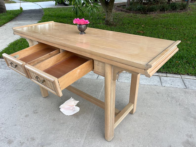 May include: A light-colored wooden console table with two drawers open. The drawers have woven rattan accents and ornate handles. A small silver vase with a pink flower sits on top. The table is approximately 60 inches long.