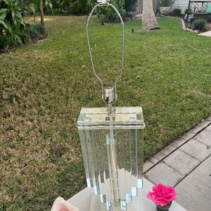 May include: A clear acrylic table lamp with a rectangular base and a series of vertical panels. The lamp has a silver-colored metal stem and a clear glass shade holder. A pink rose in a small black pot and a conch shell are on the table.