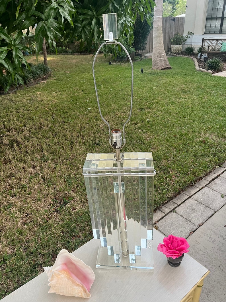 May include: A clear acrylic table lamp with a rectangular base and a series of vertical panels. The lamp has a silver-colored metal stem and a clear glass shade holder. A pink rose in a small black pot and a conch shell are on the table.