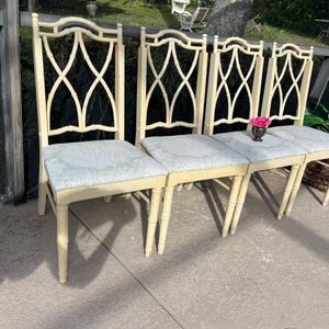 May include: Four cream-colored dining chairs with light blue patterned seats. The chairs have a decorative back with an X-shaped design. A small vase with a pink flower sits on the seats. The chairs are outdoors.