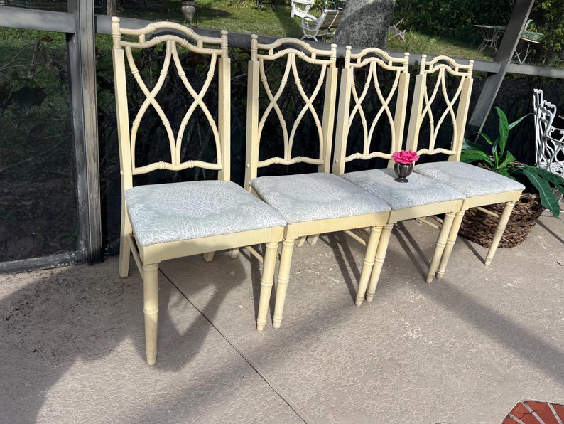 May include: Four cream-colored dining chairs with light blue patterned seats. The chairs have a decorative back with an X-shaped design. A small vase with a pink flower sits on the seats. The chairs are outdoors.