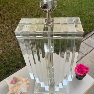 May include: A clear acrylic lamp with a rectangular, stepped base and a shade composed of vertical, clear acrylic panels. The lamp has a silver metal stem and a standard light bulb socket. A seashell and a pink rose in a small black vase are next to the lamp.