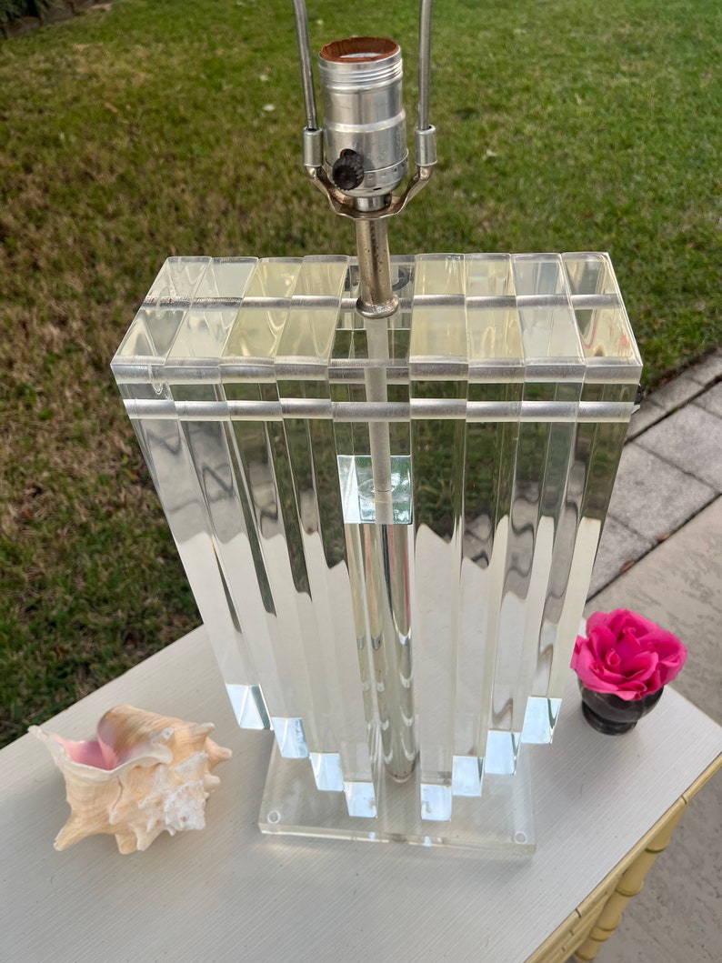 May include: A clear acrylic lamp with a rectangular, stepped base and a shade composed of vertical, clear acrylic panels. The lamp has a silver metal stem and a standard light bulb socket. A seashell and a pink rose in a small black vase are next to the lamp.