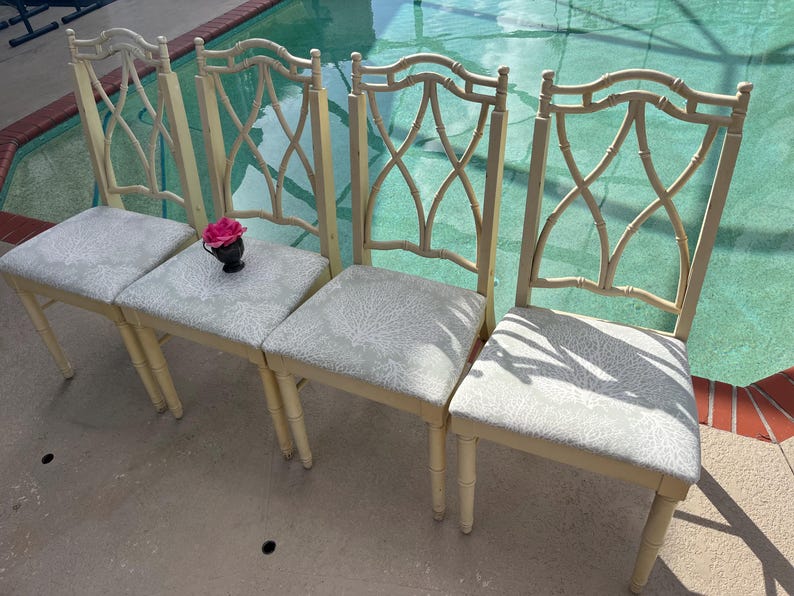 May include: Four cream-colored dining chairs with light gray patterned seats. The chair backs have an open, decorative design. A small black vase with a pink flower sits on one of the seats. The chairs are on a patio near a pool.