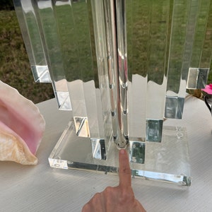May include: A clear acrylic table lamp with a geometric design. The lamp features a rectangular base and a series of vertical, rectangular panels. The lamp is next to a conch shell and a small silver vase with a pink rose.