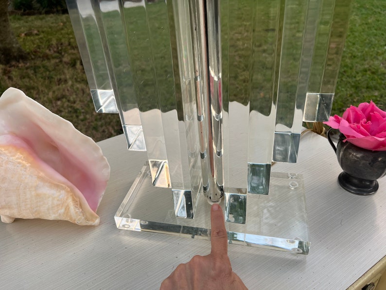 May include: A clear acrylic table lamp with a geometric design. The lamp features a rectangular base and a series of vertical, rectangular panels. The lamp is next to a conch shell and a small silver vase with a pink rose.