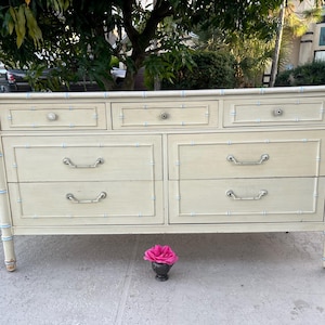 May include: A cream-colored dresser with seven drawers and silver-toned hardware. The frame has a bamboo-style design with light blue accents. A small, dark vase holds a pink rose in front of the dresser. The dresser is outdoors.