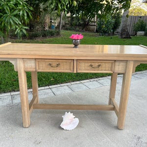 May include: A light-colored wooden console table with two drawers featuring woven rattan fronts and brass handles. A small silver vase with pink flowers sits on top. A large seashell rests on the ground in front of the table.