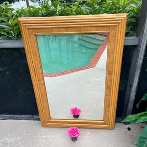 May include: A rectangular mirror with a light brown bamboo frame. The mirror reflects a pool with turquoise water and a red brick border. Two pink flowers sit in front of the mirror.