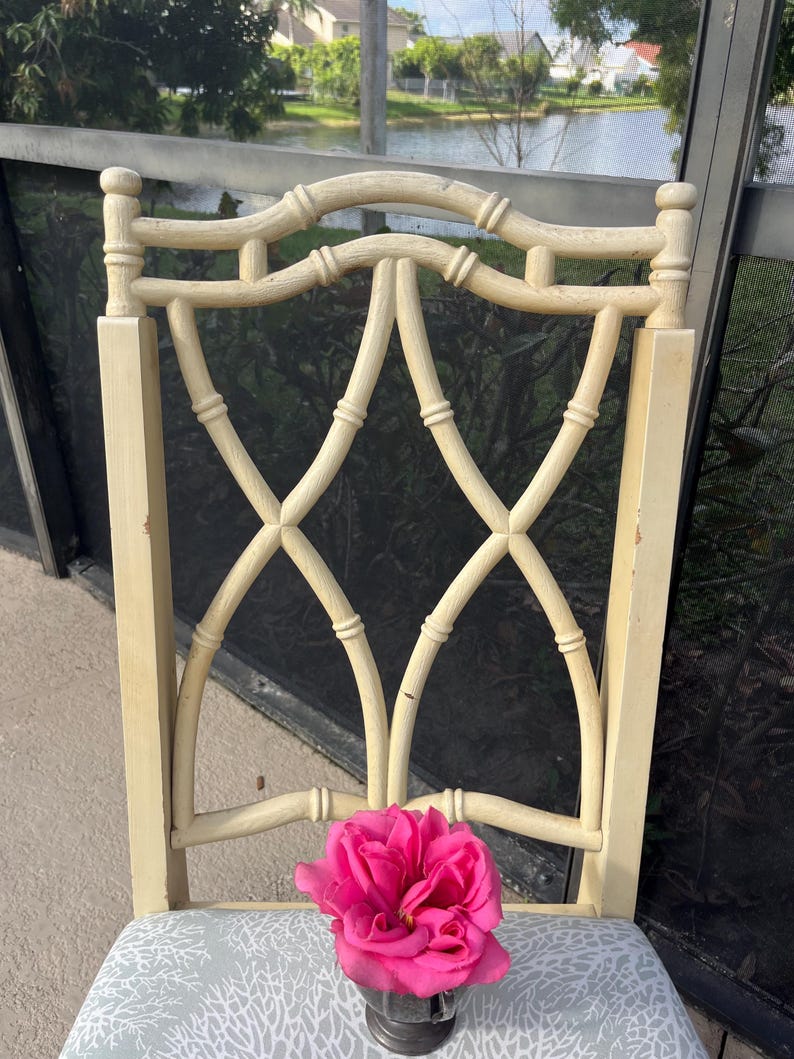 May include: A cream-colored wooden chair with a decorative back featuring a lattice design. The chair has a patterned seat cushion and a vibrant pink rose in a small metal container resting on the seat.