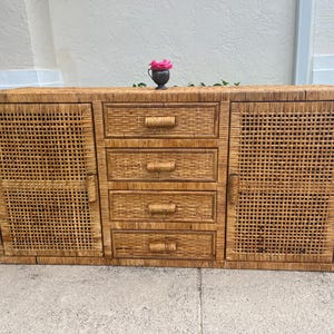 May include: A large, rectangular, light brown woven rattan cabinet with four drawers and two doors. The cabinet has a natural finish and a decorative woven pattern. A small, dark vase with a pink flower sits on top.