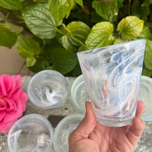 May include: Set of four clear glass cups with a swirling white pattern. The cups are arranged on a table with a pink rose in the foreground.