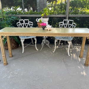 May include: A rectangular, light-colored wooden table with ornate carvings along the edges and legs. Two white metal chairs with decorative backs are positioned on either side. A seashell and flowers adorn the table's surface.