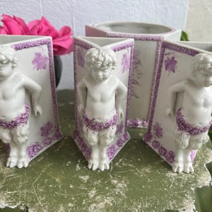 May include: A collection of white ceramic vases with a classical design. Each vase features a sculpted figure of a cherub with purple floral accents. The vases are arranged on a distressed green surface, with pink flowers in the background.