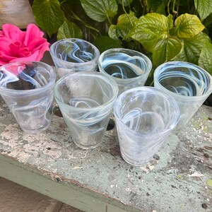 May include: Six clear glass tumblers with a swirling white pattern. The glasses are arranged in a circle on a weathered wooden surface.