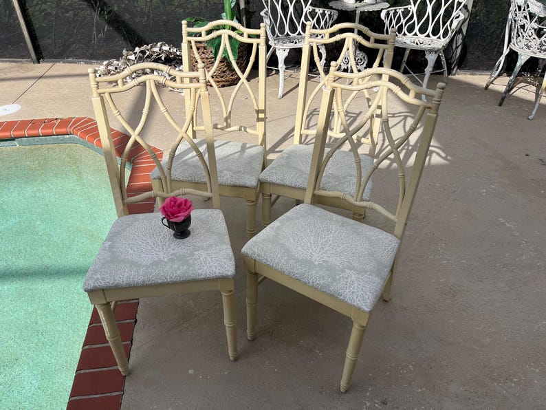 May include: Four cream-colored dining chairs with light blue patterned seats. The chairs have a decorative back design and are positioned near a pool. A small black cup with a pink flower sits on one chair.