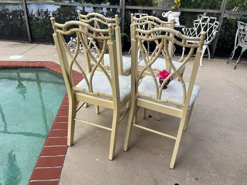 May include: Four cream-colored bamboo-style chairs with patterned seat cushions. The chairs have a decorative back design and are arranged outdoors near a pool. A pink flower is placed on one of the chairs.
