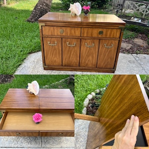 May include: A wooden cabinet with a warm brown finish, featuring four doors with brass handles and two drawers. The cabinet has brass corner accents and a pull-out top. A seashell and a small vase with flowers are on top.