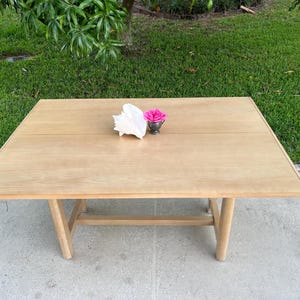 May include: A light-colored wooden table with a rectangular top and angled legs. A white seashell and a small silver cup with a pink flower are on the table's surface. The table is outdoors on a concrete surface.
