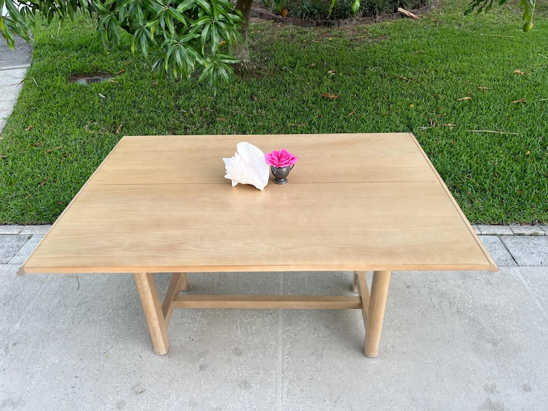 May include: A light-colored wooden table with a rectangular top and angled legs. A white seashell and a small silver cup with a pink flower are on the table's surface. The table is outdoors on a concrete surface.