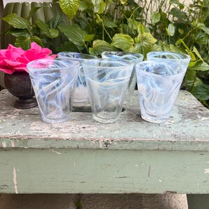 May include: Five clear glass tumblers with a white swirl design. The glasses are sitting on a green wooden surface.