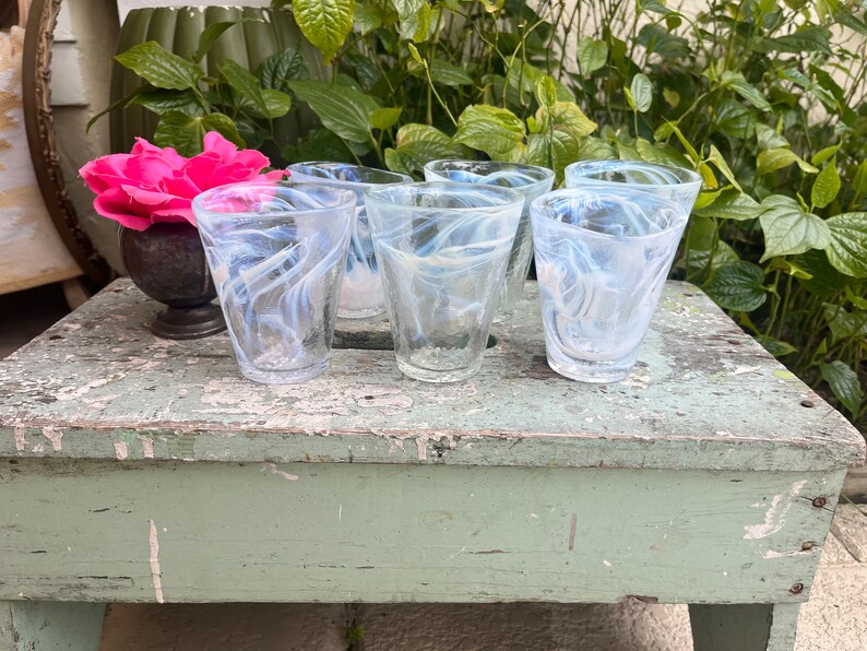 May include: Five clear glass tumblers with a white swirl design. The glasses are sitting on a green wooden surface.