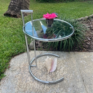 May include: A chrome and glass side table with a round top and a single shelf. The table has a curved base and a handle on the side. A pink flower is in a vase on the table.