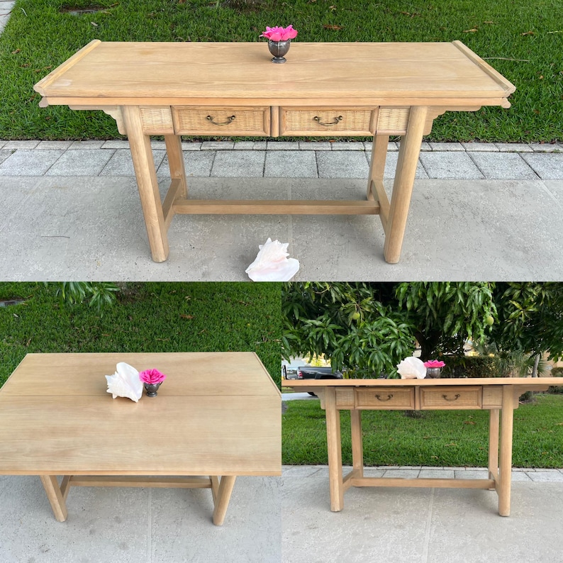 May include: A light-colored wooden desk with two drawers featuring woven rattan fronts. The desk has a rectangular top and angled legs. A small silver vase with pink flowers sits on top. A seashell is also present.